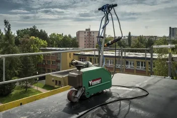 Svařovací automat Leister Varimat na ploché střeše, v pozadí panelové domy a škola.