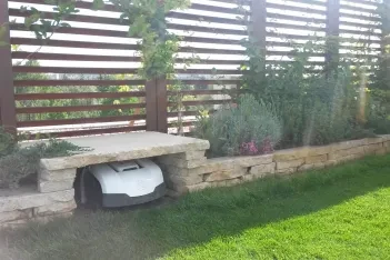 Robotická sekačka ve stanici pod kamennou lavicí u trávníku na zelené střeše.